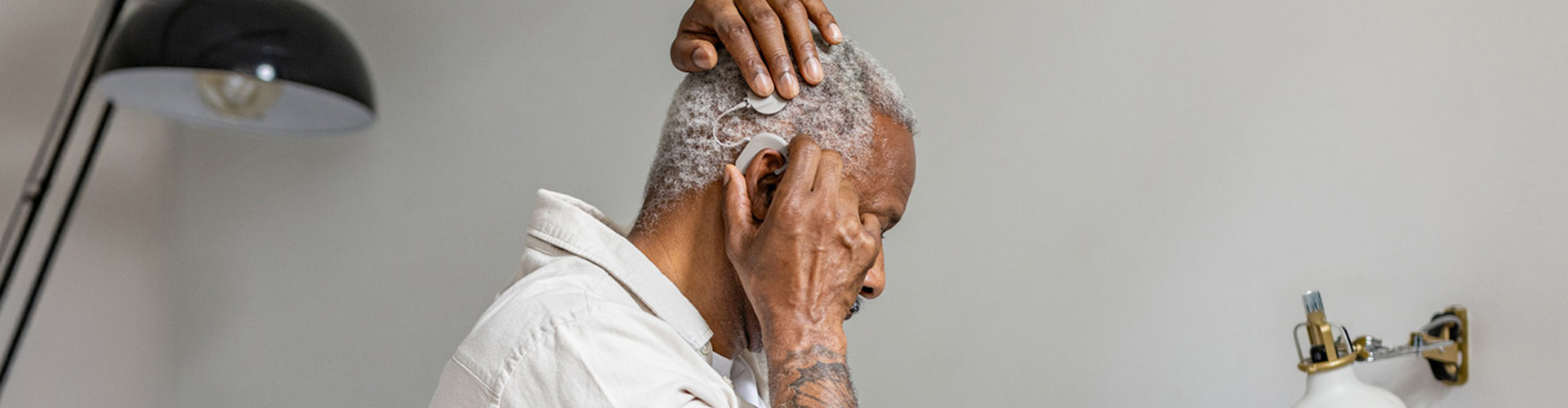 An older man puts on his SONNET 3 cochlear implant audio processor while indoors near a lamp.