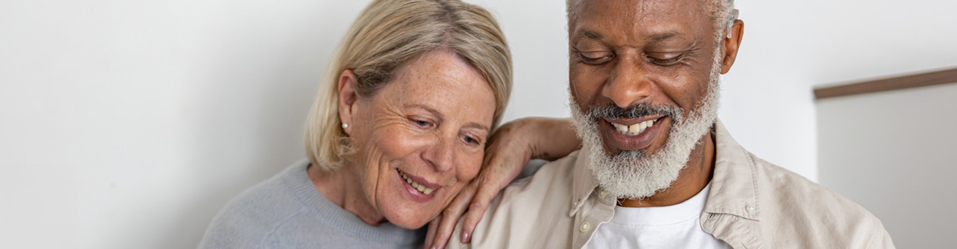 A couple sit close together at home. The man has a cochlear implant audio processor behind his ear.