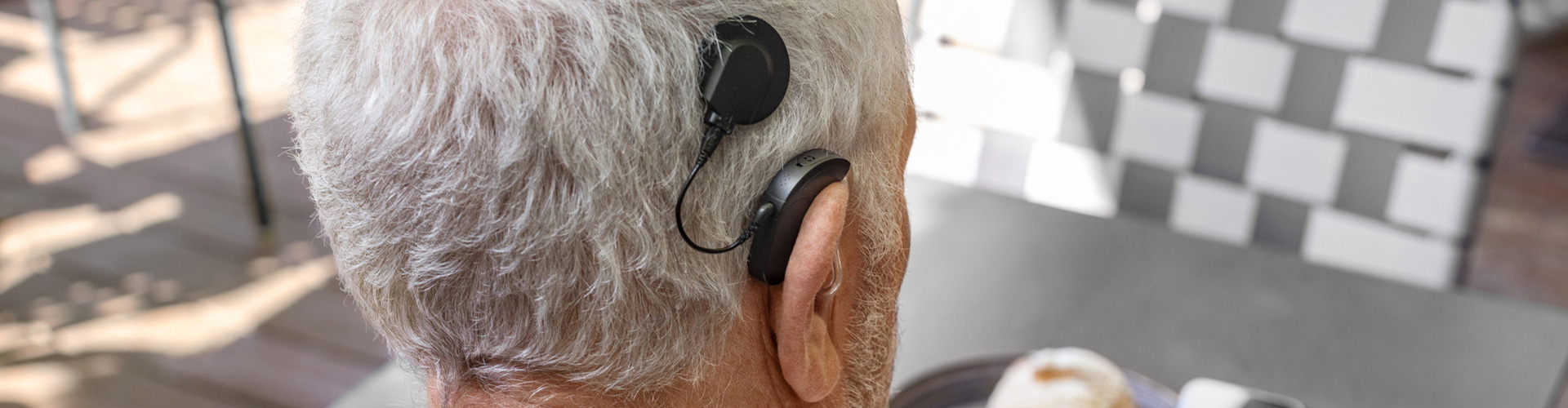 Close-up of the back of a person’s head with short gray hair, showing a black cochlear implant audio processor. The setting includes an outdoor café table with a plate and cup partially visible.