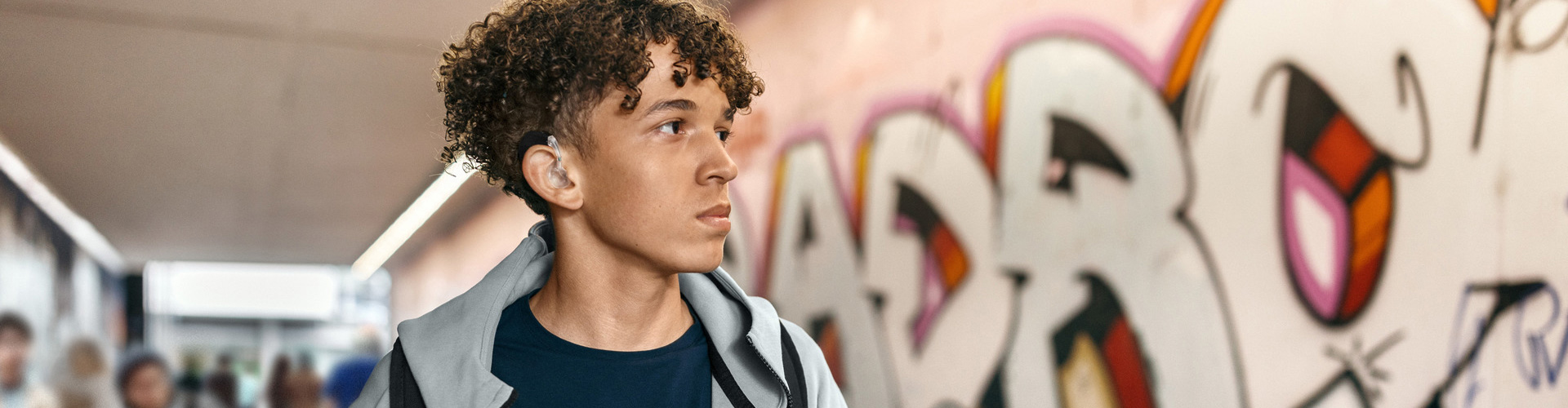Young man wearing a cochlear implant audio processor walking through an urban tunnel with colorful graffiti art on the walls. Bright daylight is visible at the tunnel exit.